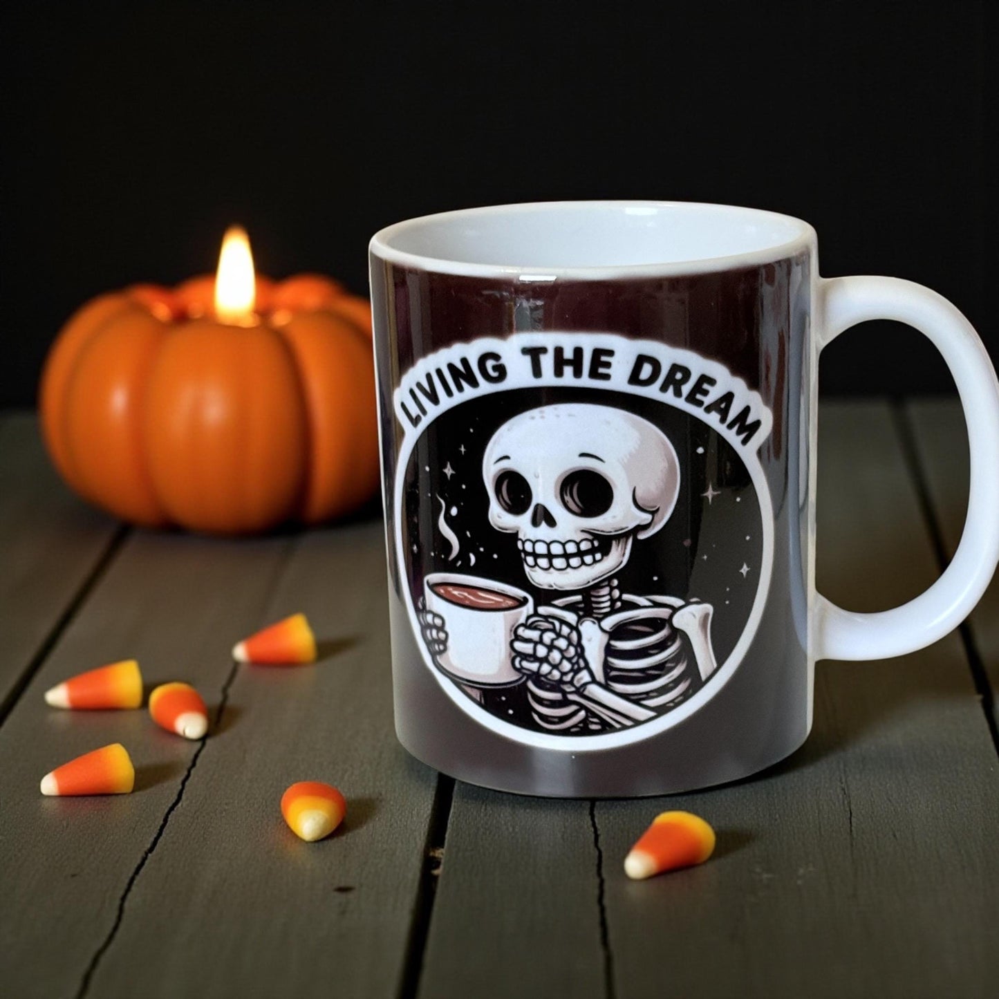 Mug with skeleton design and 'Living the Dream' text, surrounded by pumpkins and candy corn on a wooden surface.