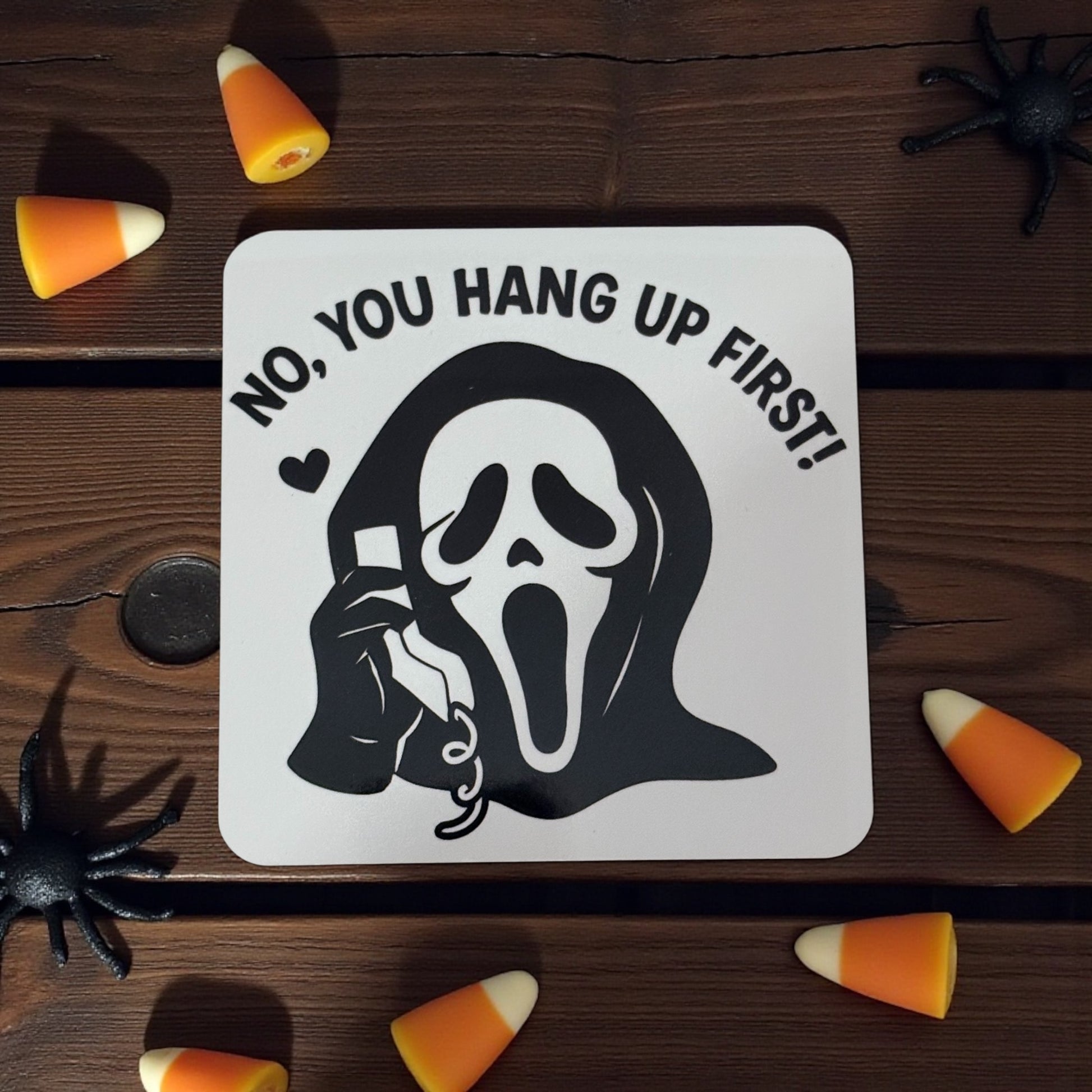 Halloween-themed coaster with ghostly design and text on a wooden surface with candy corn and spiders.