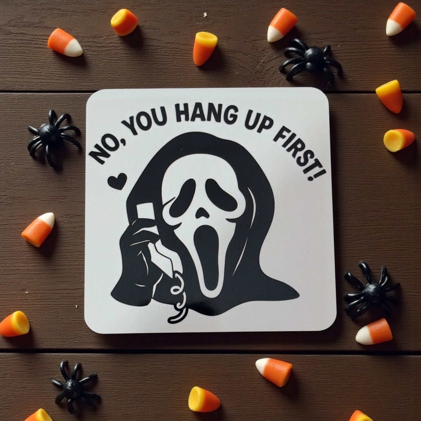 Halloween-themed coaster with ghost and text 'No, you hang up first!' surrounded by candy corn and spiders on a wooden surface.