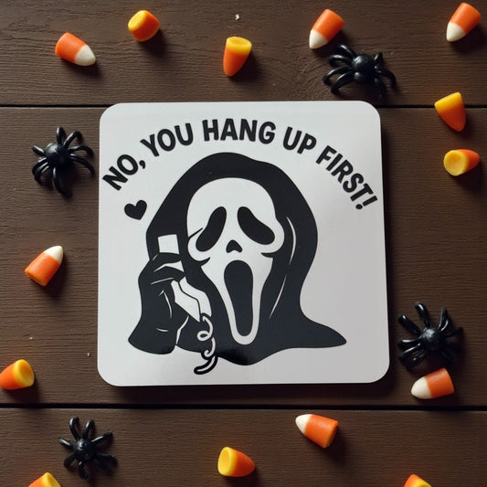 Halloween-themed coaster with ghost and text 'No, you hang up first!' surrounded by candy corn and spiders on a wooden surface.