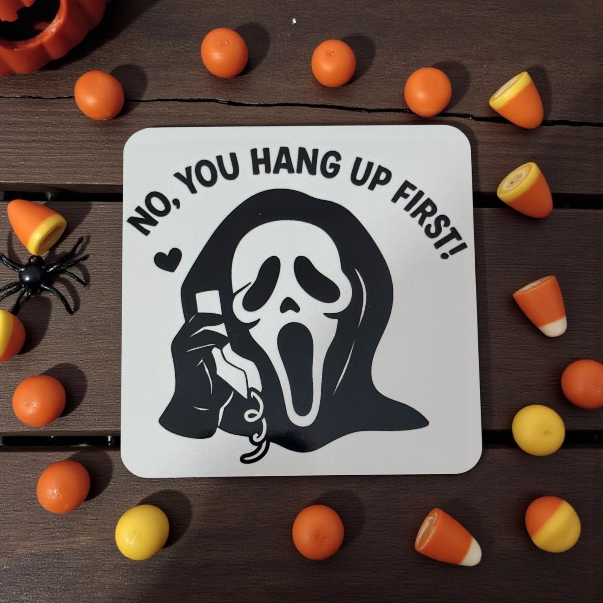 Halloween-themed coaster with ghost and text 'No, you hang up first!' surrounded by candy on a wooden surface.