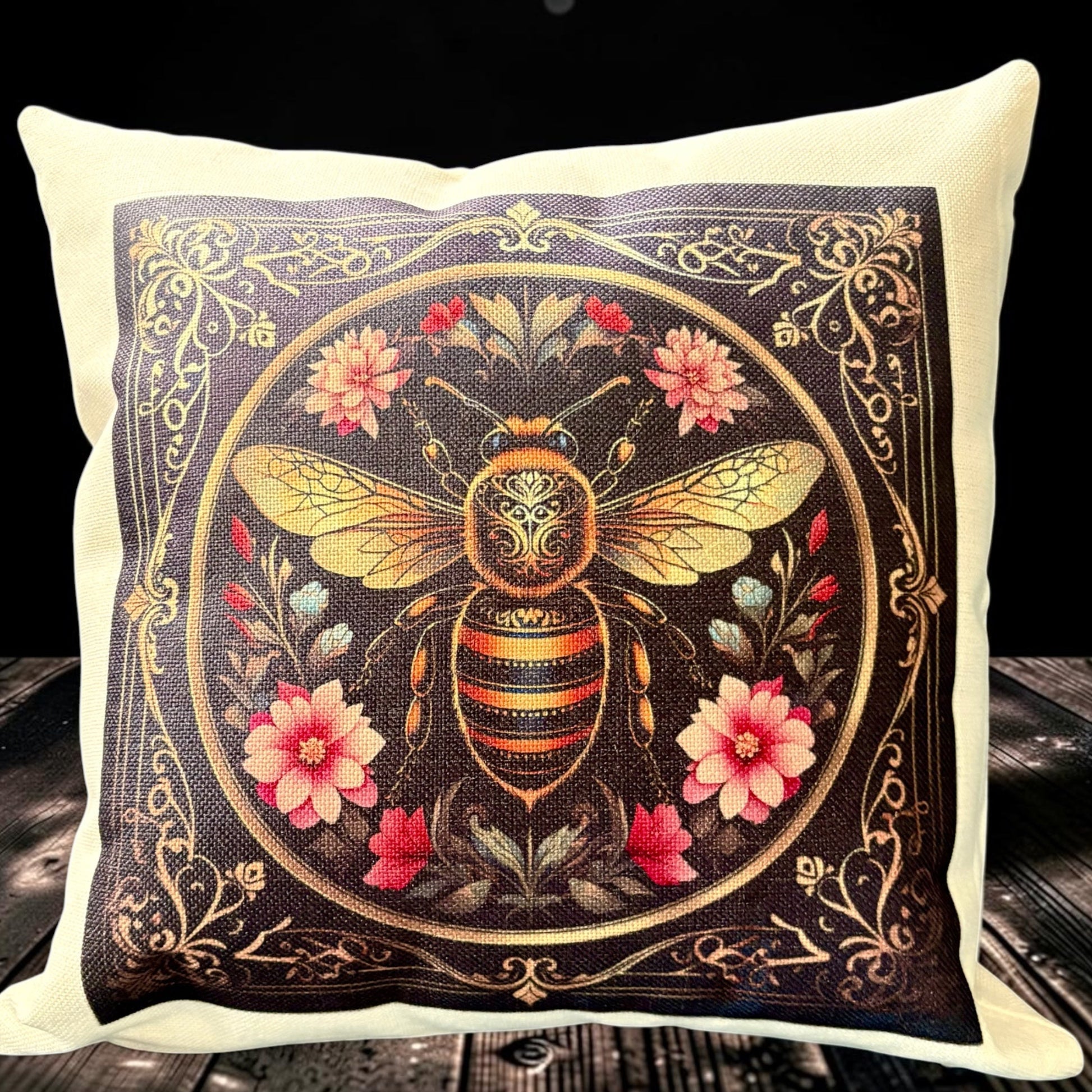 A square throw pillow with a cream-colored fabric border and a central printed design of a detailed golden bee surrounded by pink and red flowers on a dark background, displayed on a rustic wooden surface against a black backdrop.