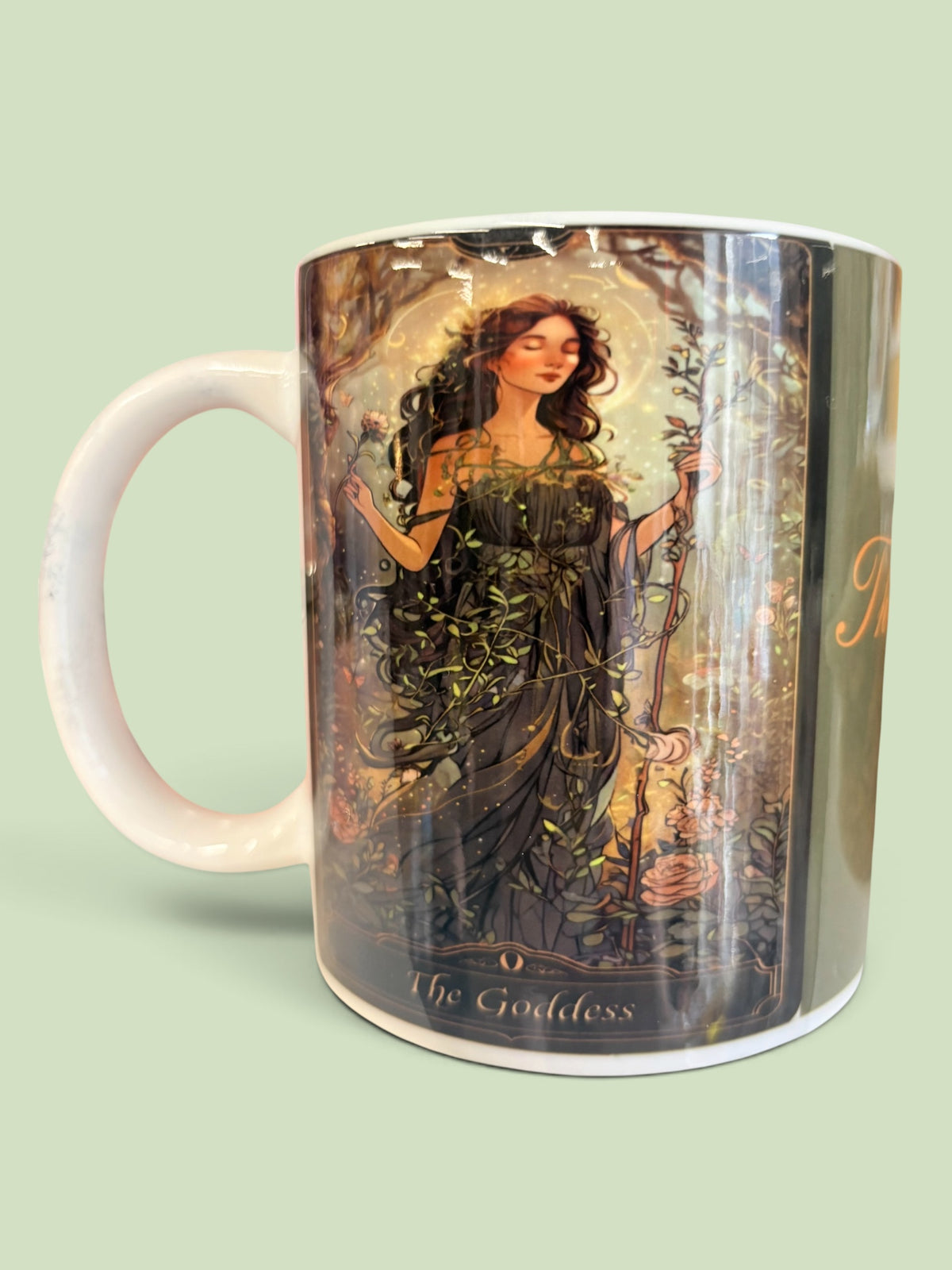 A white ceramic mug featuring a detailed fantasy illustration of 'The Goddess' tarot card character, set against a solid pale green background.