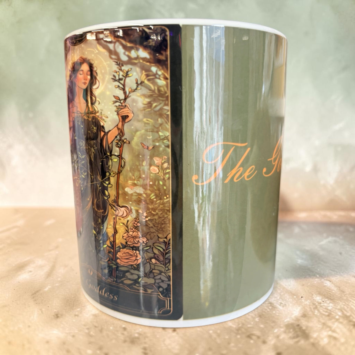 A ceramic mug featuring the 'The Goddess' tarot card design, showing a dark green side with gold cursive text and a detailed illustration of a goddess holding a staff on the other side.