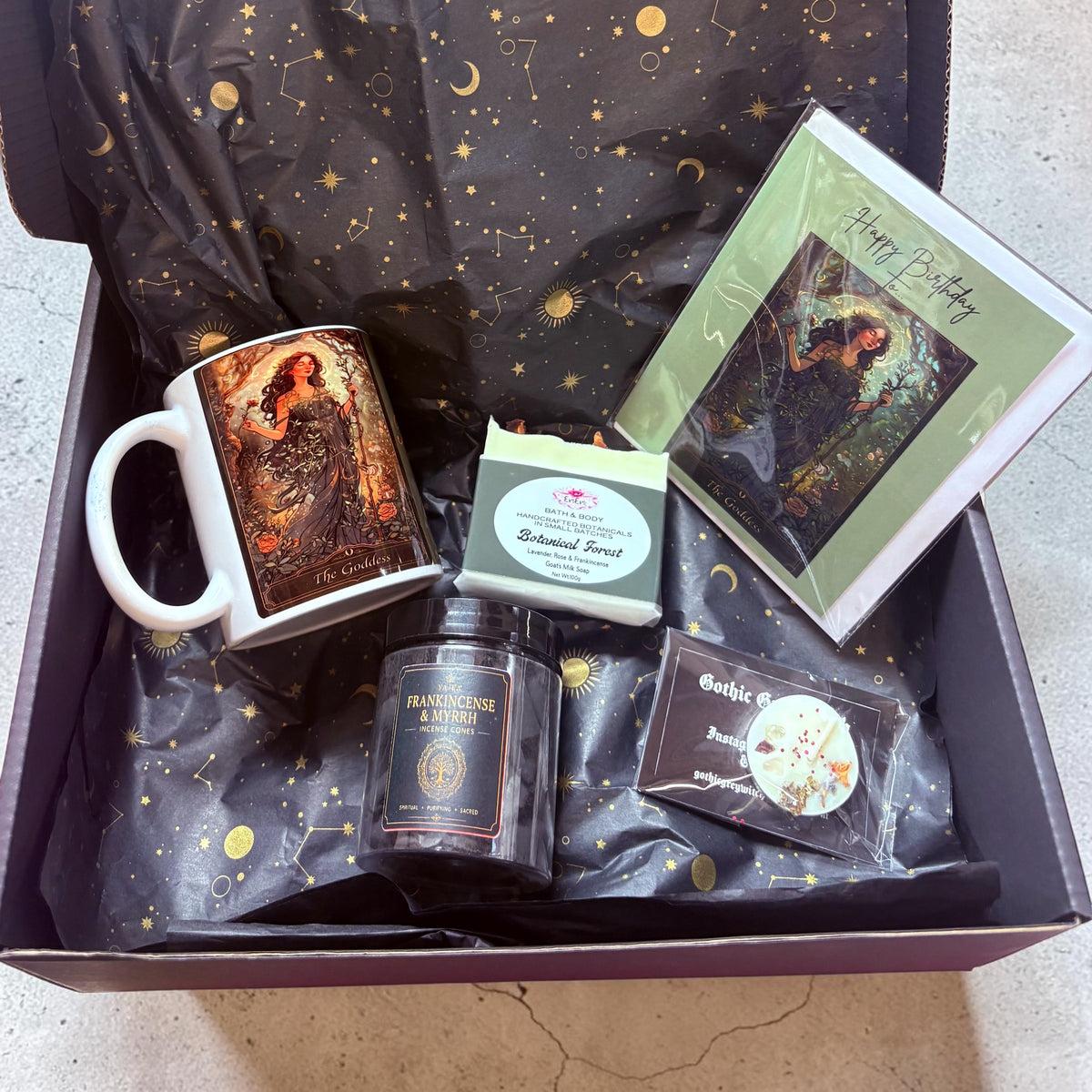 A curated gift box filled with fantasy-themed items, including a white ceramic mug with an illustration of The Goddess in a flowing dress, a tea light candle with rose chrystals, a small handmade soap bar, and a Goddess card, all arranged on black starry tissue paper and enclosed in a black gift box