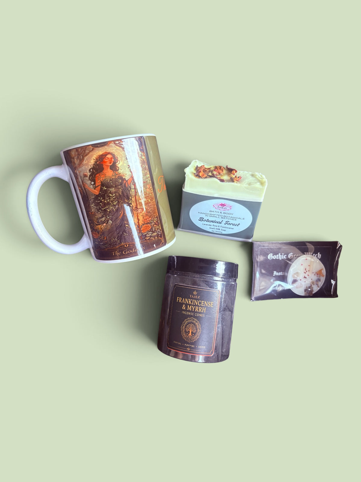A curated flat-lay of four distinct products on a solid pale sage green background: a white ceramic mug with a vintage illustration, a bar of soap labeled 'Botanical Forest" a dark brown incense tin labeled 'Frankencake & Myrh, and a small packaged rose quartz tea light