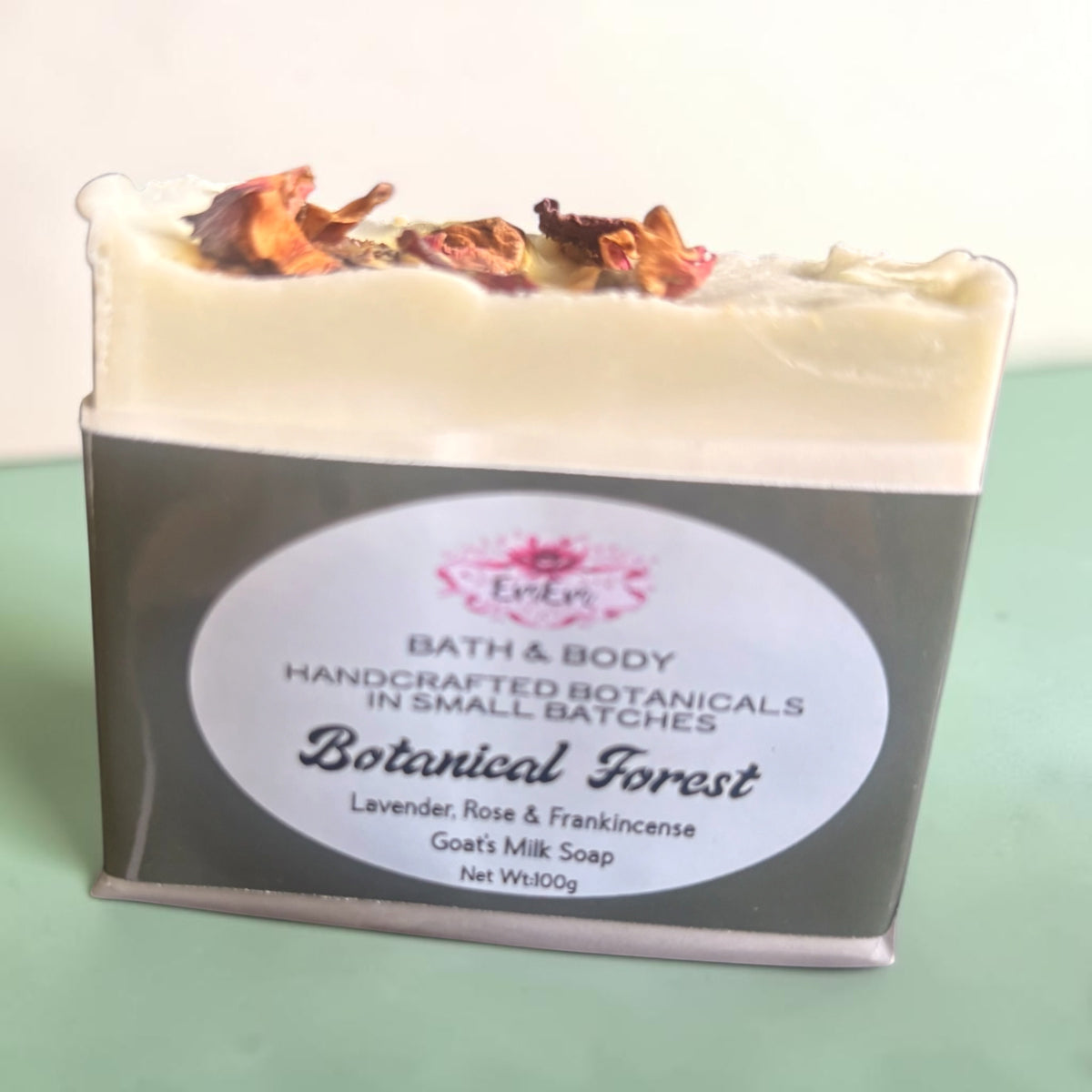 A rectangular bar of handmade bath & body soap labeled 'Botanical Forest' by 'EmEm Bath & Body sits on a pale green surface against a white background. The Goat's Milk soap has a cream-colored top layer with dried botanical pieces, scented with Lavender, Frankincense and Rose wrapped in forest green packaging
