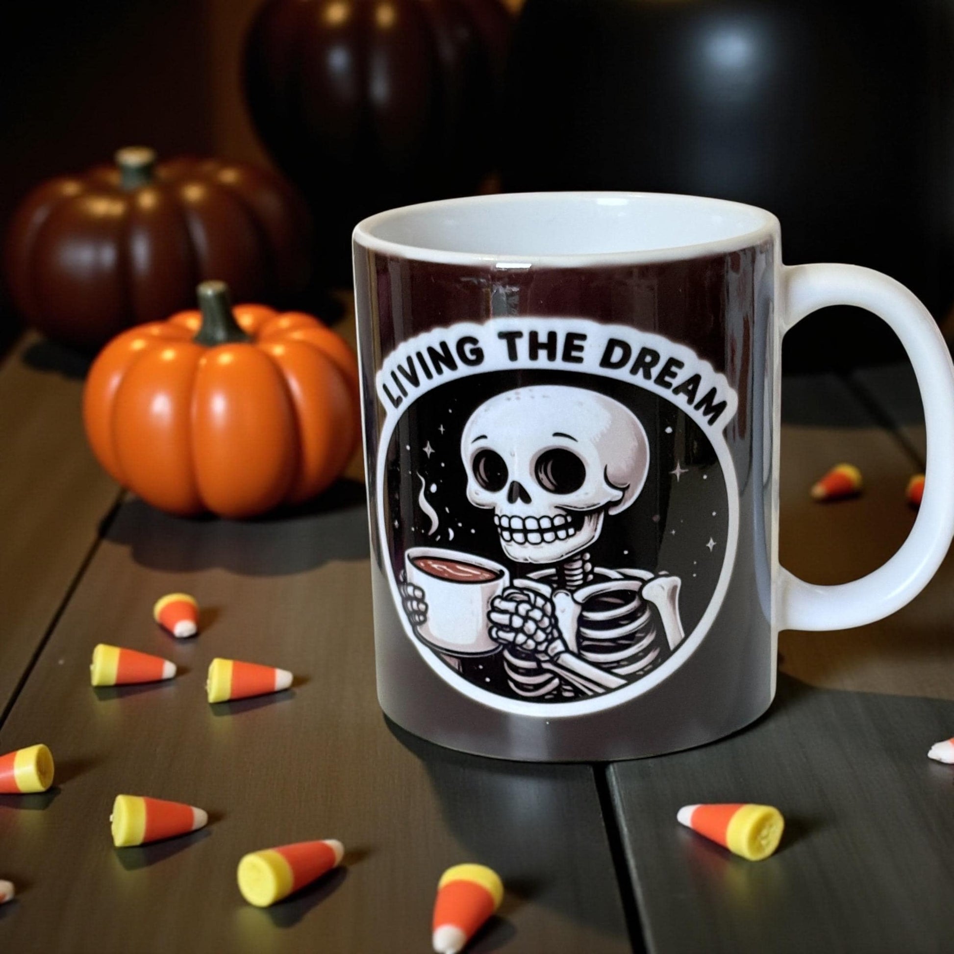 Mug with skeleton design and 'Living the Dream' text, surrounded by pumpkins and candy corn on a wooden surface.