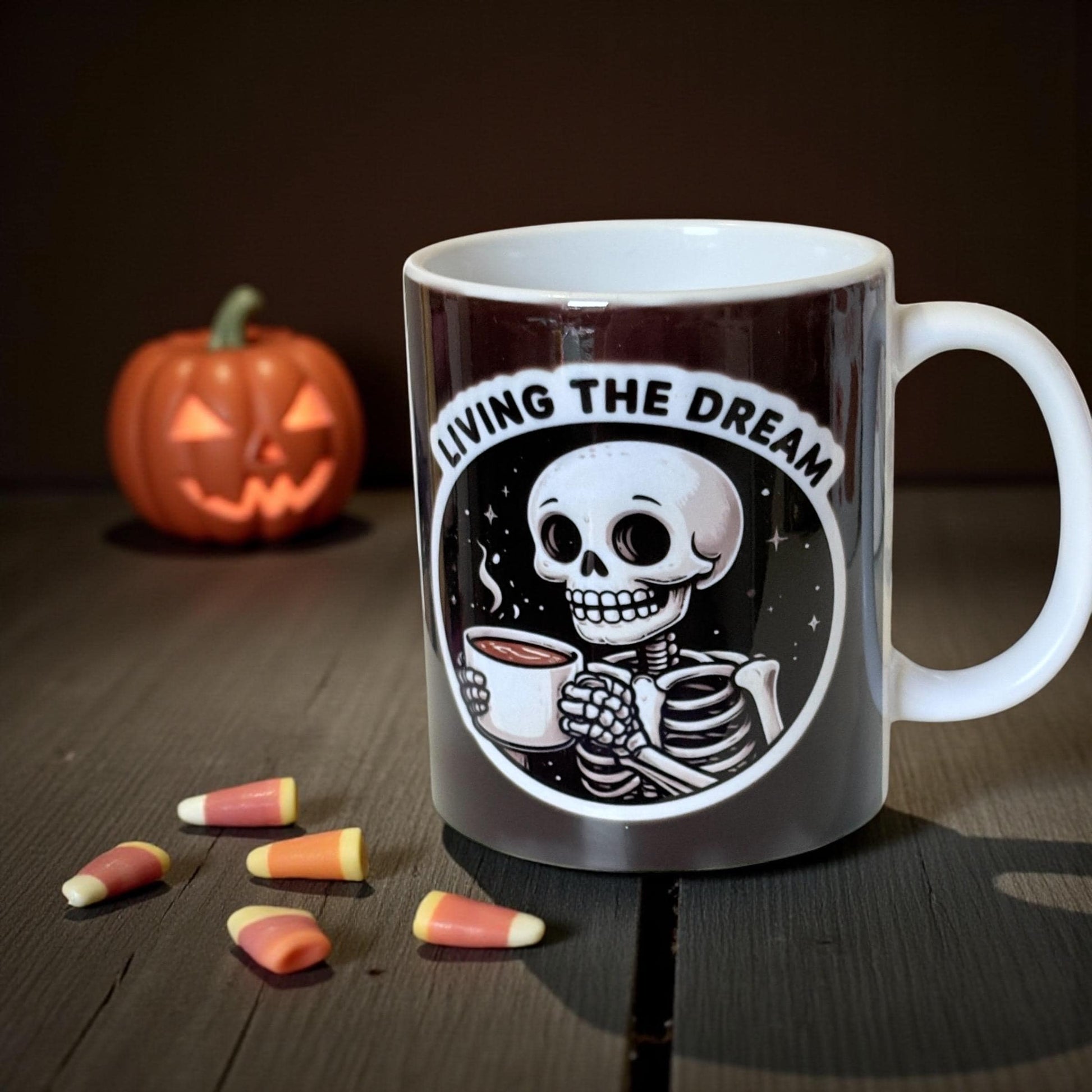 Mug with skeleton design and 'Living the Dream' text, surrounded by candy corn and a pumpkin on a wooden surface.
