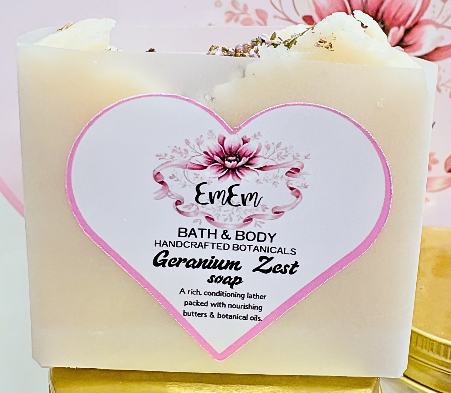 A square bar of EmEm Bath & Body soap labeled 'Geranium Zest soap' in a translucent cream-colored container with a large white heart-shaped label featuring pink accents and botanical illustrations.